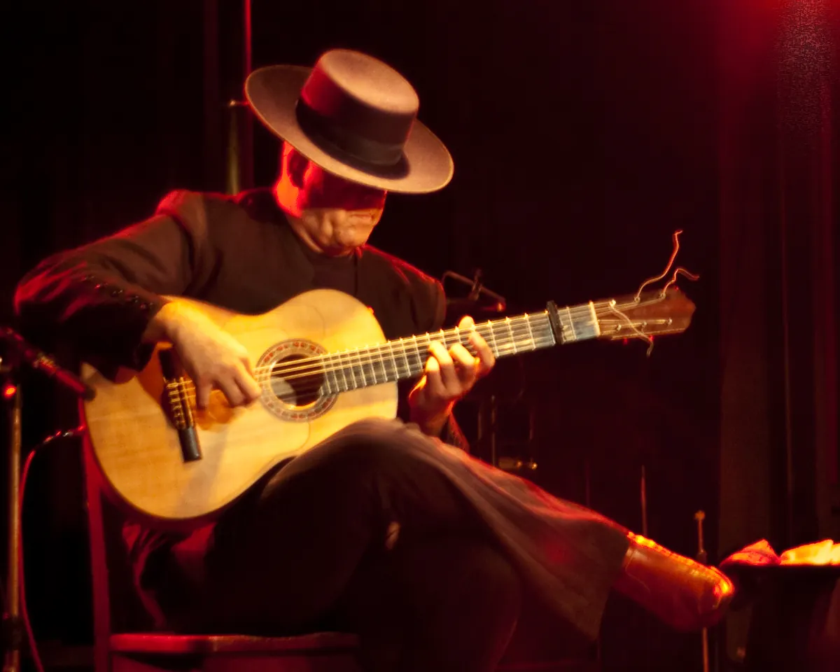 Flamenco kytarista v širokém klobouku hrající na světlou kytaru v červeném jevištním světle