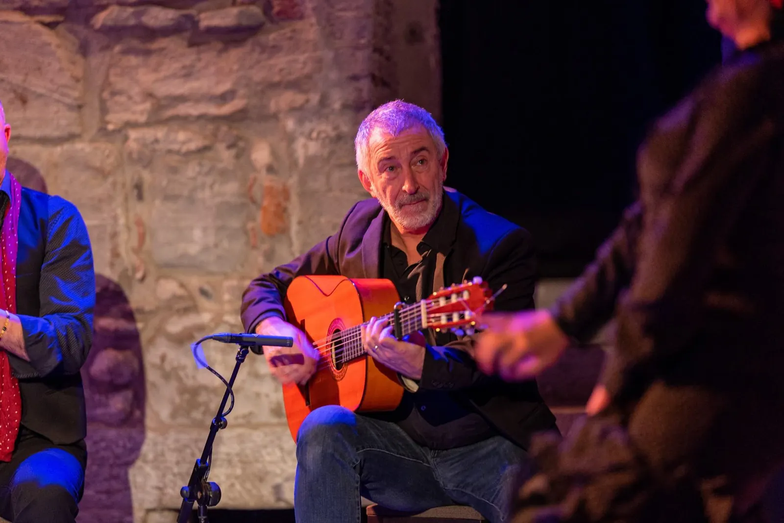 En flamencoguitarist i mørk jakke, der spiller under scenelys ved en stenmur, med en anden udøver, der klapper ved siden af ham