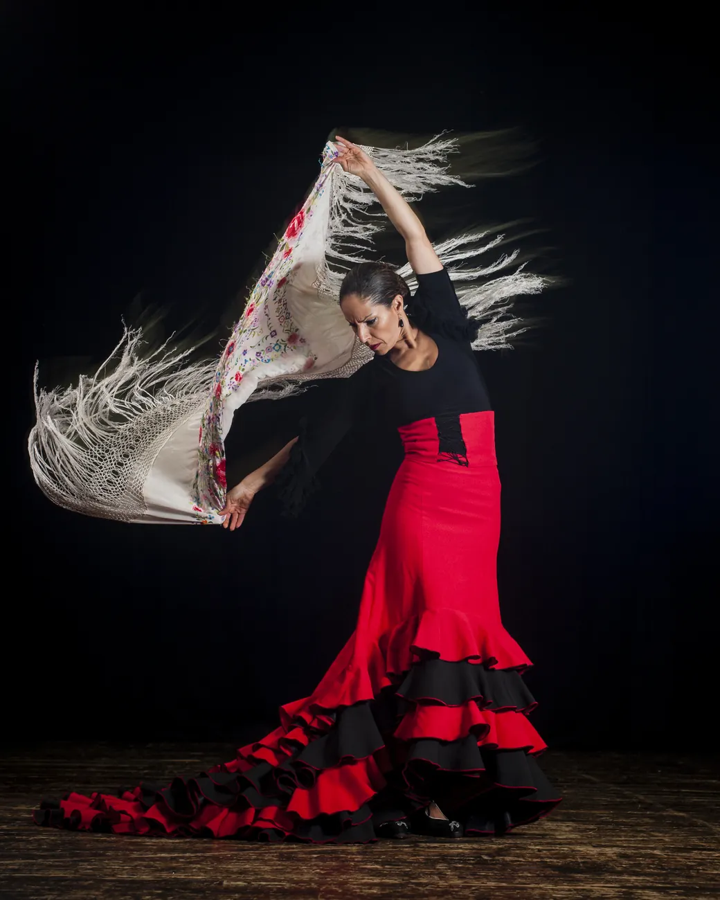 Flamenková tanečnice v černých a červených šatech rozvíjející šál s třásněmi na pozadí černé scény